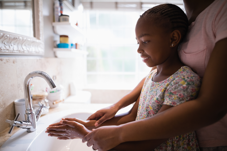 A young girl washes her hands at a sink. A grown-up helps.