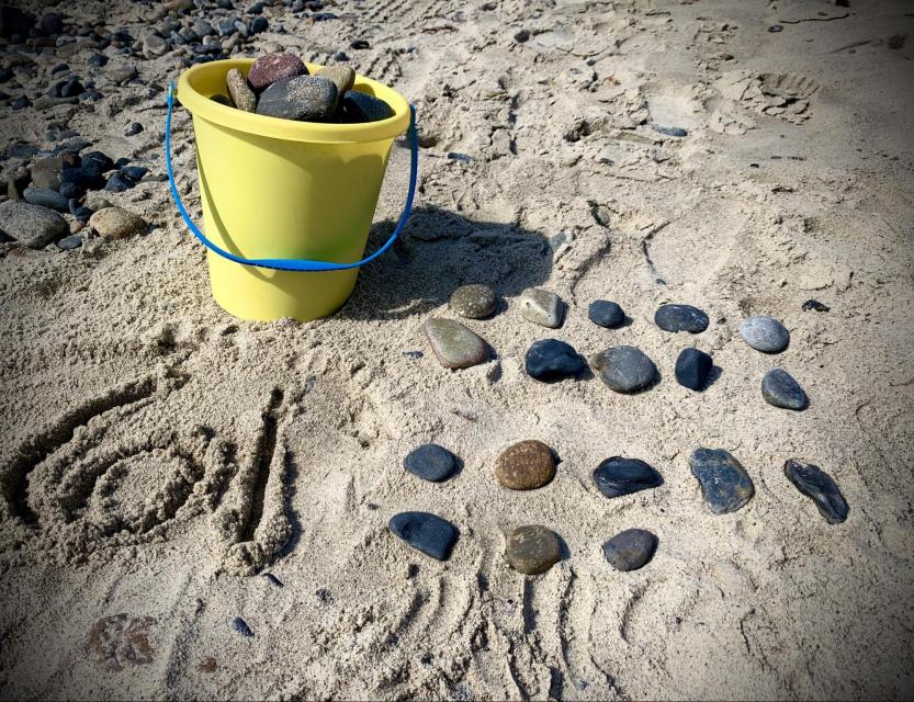 A bucket filled with rocks sits on the beach. Someone wrote 61 in the sand in front of the bucket. Beside the bucket, they arranged more rocks in 4 rows. Three rows have 5 rocks and one row has 3 rocks.