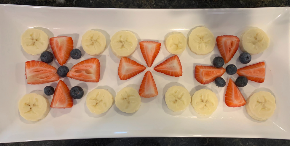 Cut up and whole fruit make 3 designs in a row. Each design has 4 slices of banana at the corners. Strawberry pieces make a plus sign between the banana slices. The strawberries might be 1-fourths. The 2 outer designs also have 5 blueberries , 1 right in the middle and 4 more in the spaces between strawberry pieces.