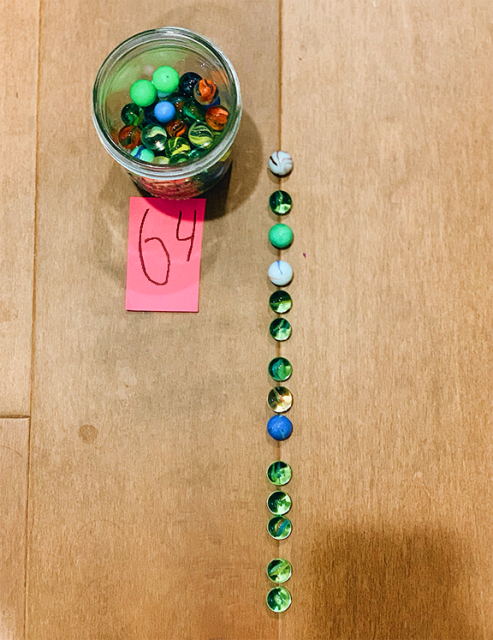 Looking down on a jar with a bunch of marbles in it. A slip of paper next to the jar has the number 64 written on it. Nearby, a line of marbles stretches down the space between two floorboards. The line looks like it has 4 sets of 3 marbles plus 2 more. The marbles have green and red and blue colors. Some colors are swirled and some are solid.