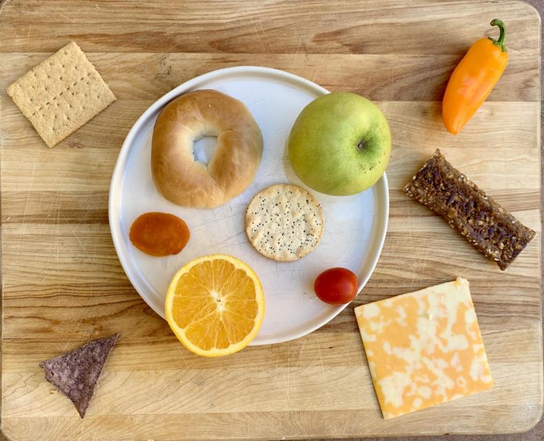 A white plate is the circle. Snacks on the plate: A bagel. An apple. 2 tomatoes. A round cracker. And a round orange slice. The snacks not on the plate: A graham cracker square. A mini bell pepper. A fruit bar rectangle. A cheese square. And a tortilla chip triangle.